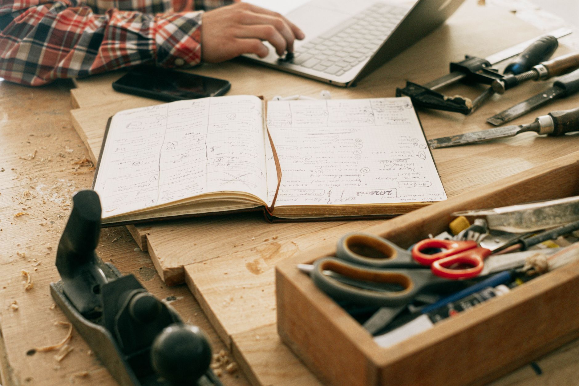 Secretária de um artesão com ferramentas, um caderno de notas e um portátil, mostrando um espaço de trabalho criativo em casa.
